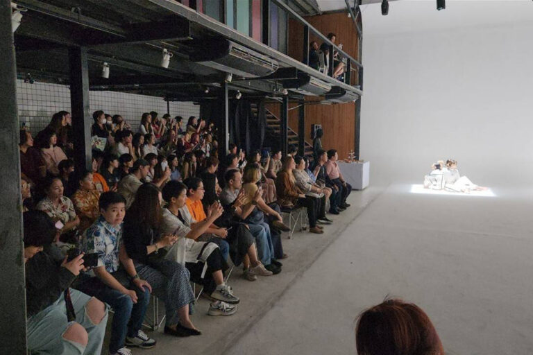 ผู้ชมนั่งเรียงแถวดูนักแสดงใต้สปอตไลต์บนพื้นขาวใน The Circus Studio