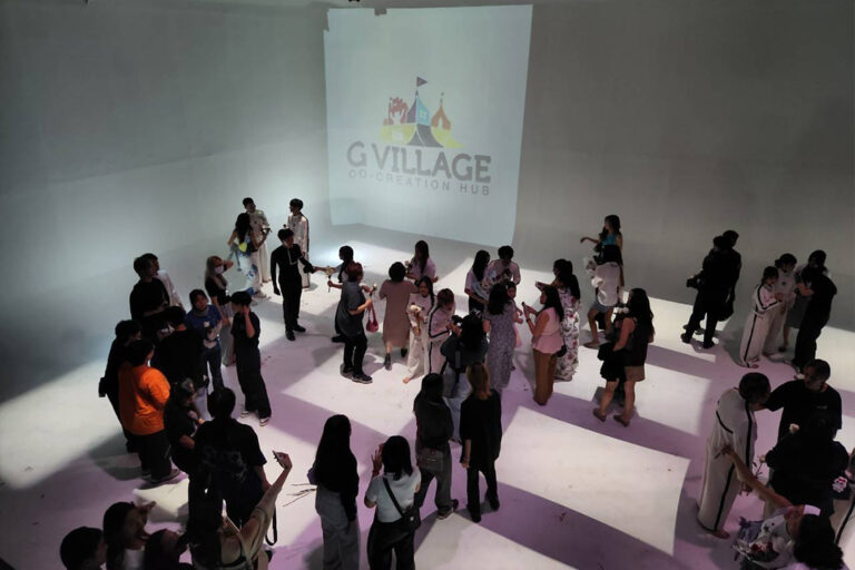 ผู้ร่วมงานเดินชมงานกลางฮอลล์ขาวที่ฉายโลโก้ G Village บนผนัง The Circus Studio