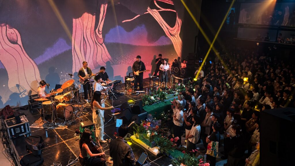วงดนตรีแสดงบนเวทีพร้อมภาพฉายและผู้ชมหน้าเวที